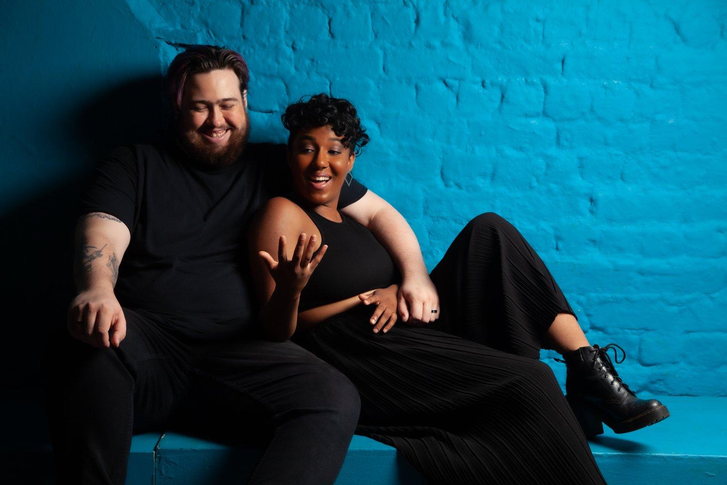 Rhyana and Matt sat against a blue brick background, mid conversation.