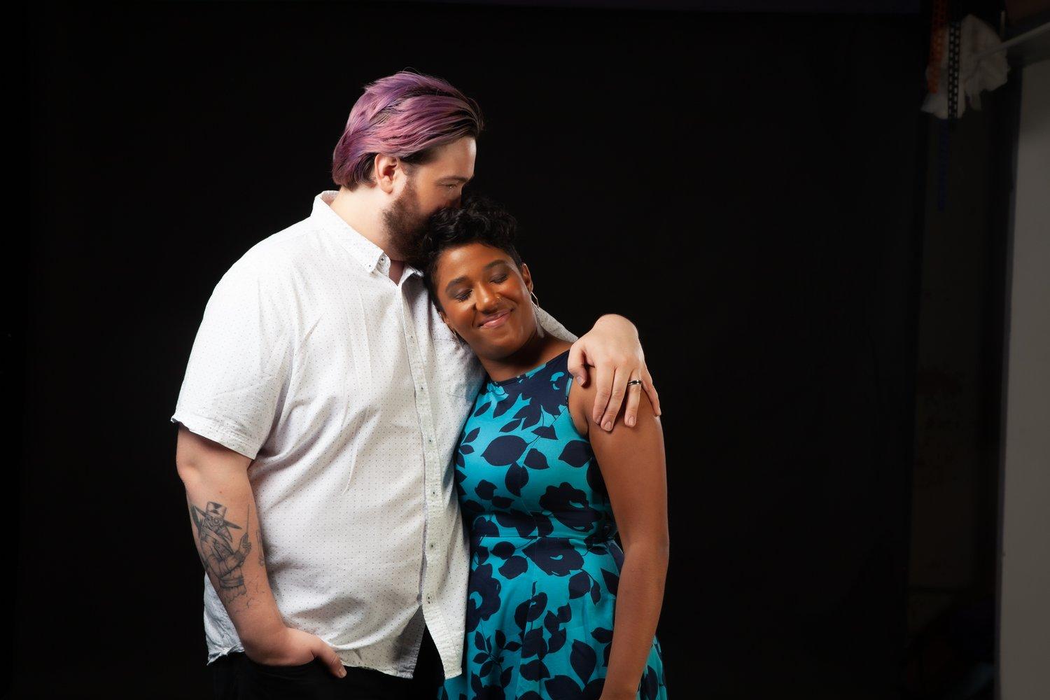 Matt kissing Rhyana on the head as they embrace against a black background.