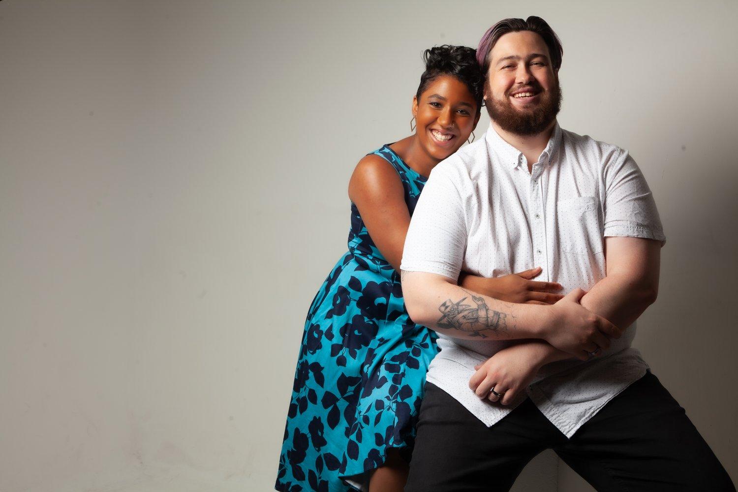 Matt sat in a white shirt while Rhyana leans against him, both smiling towards camera.