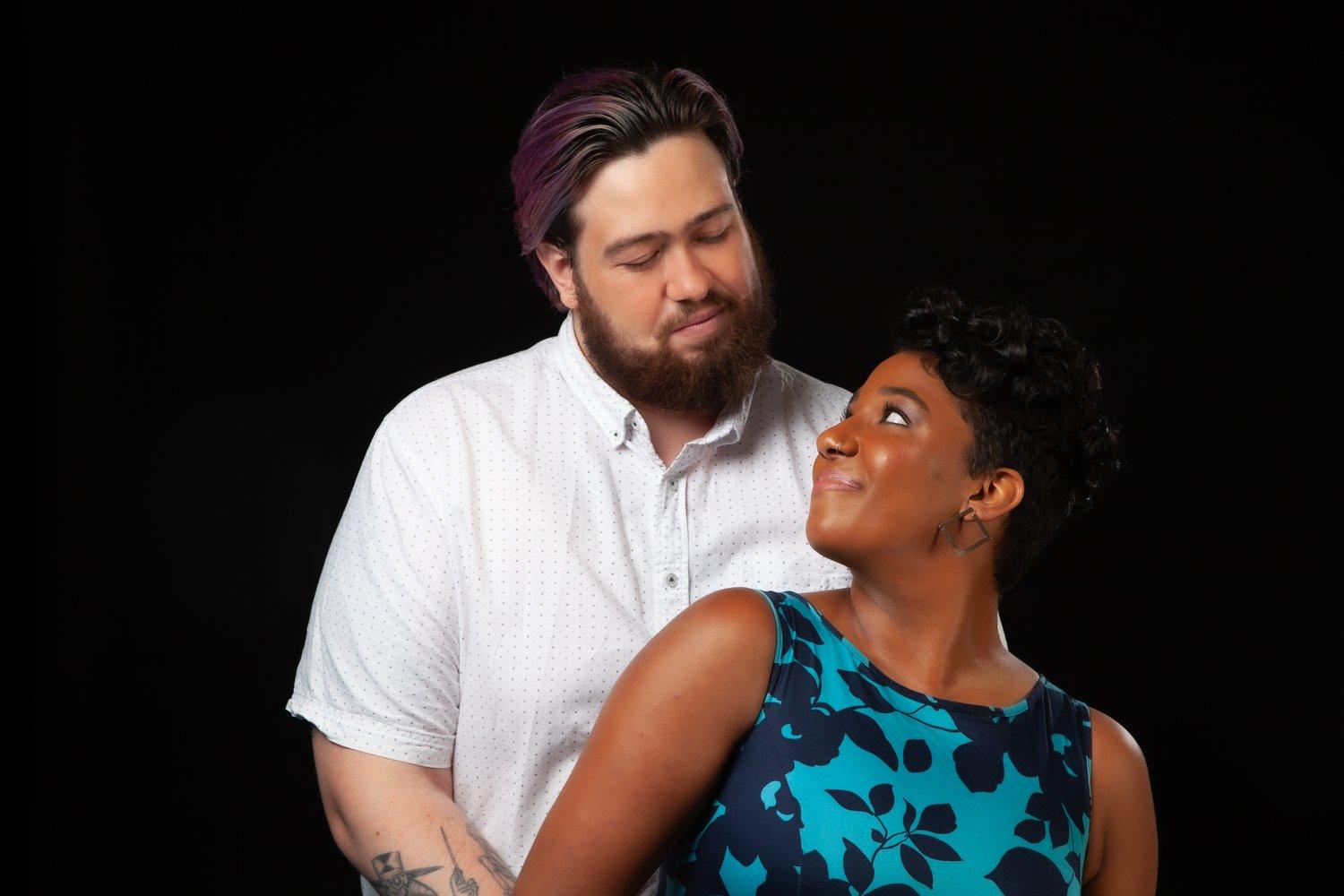 Rhyana and Matt holding hands and looking towards each other against a black background.