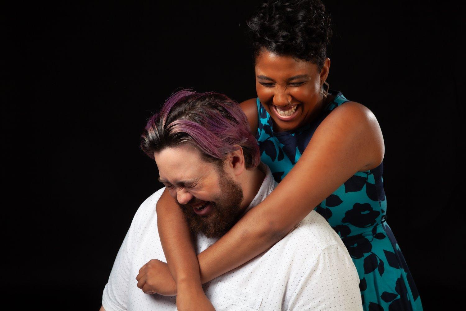 Rhyana embracing Matt from behind, who is sat in a white shirt while both laugh together.