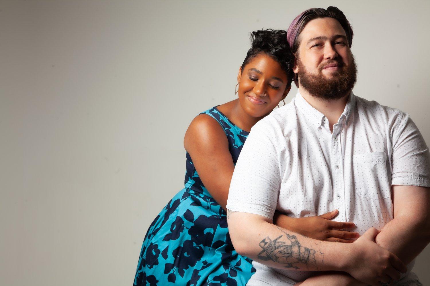 Matt sat in a white shirt while Rhyana embraces him, eyes closed.