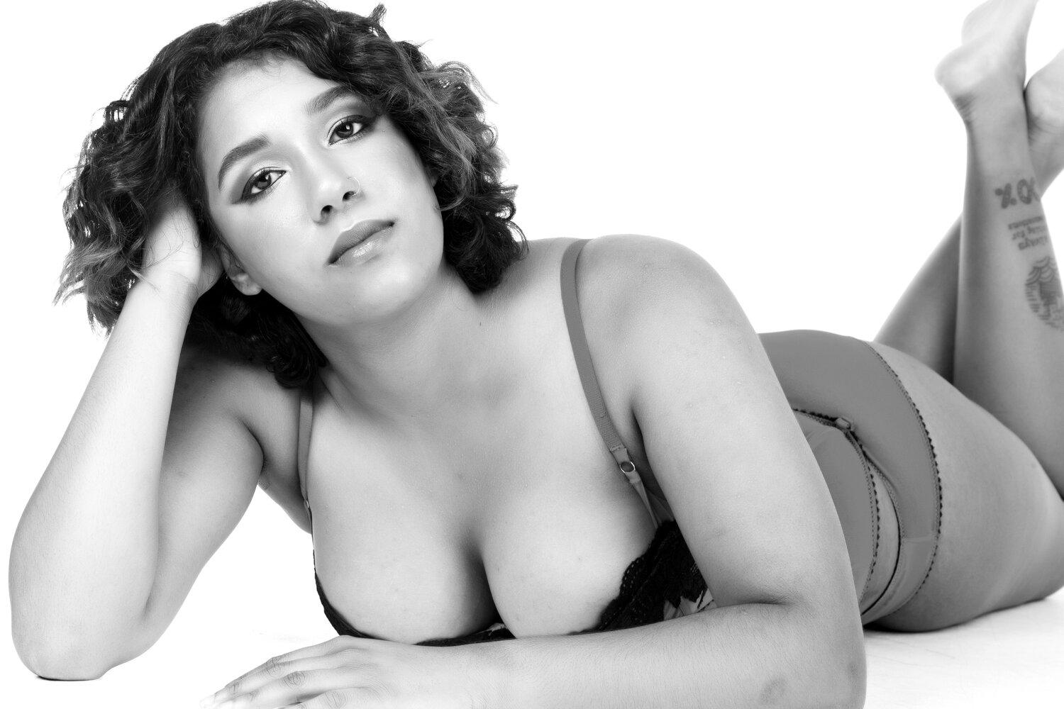 Rhyana in black and white, laying on their front up on one elbow, looking towards the camera.