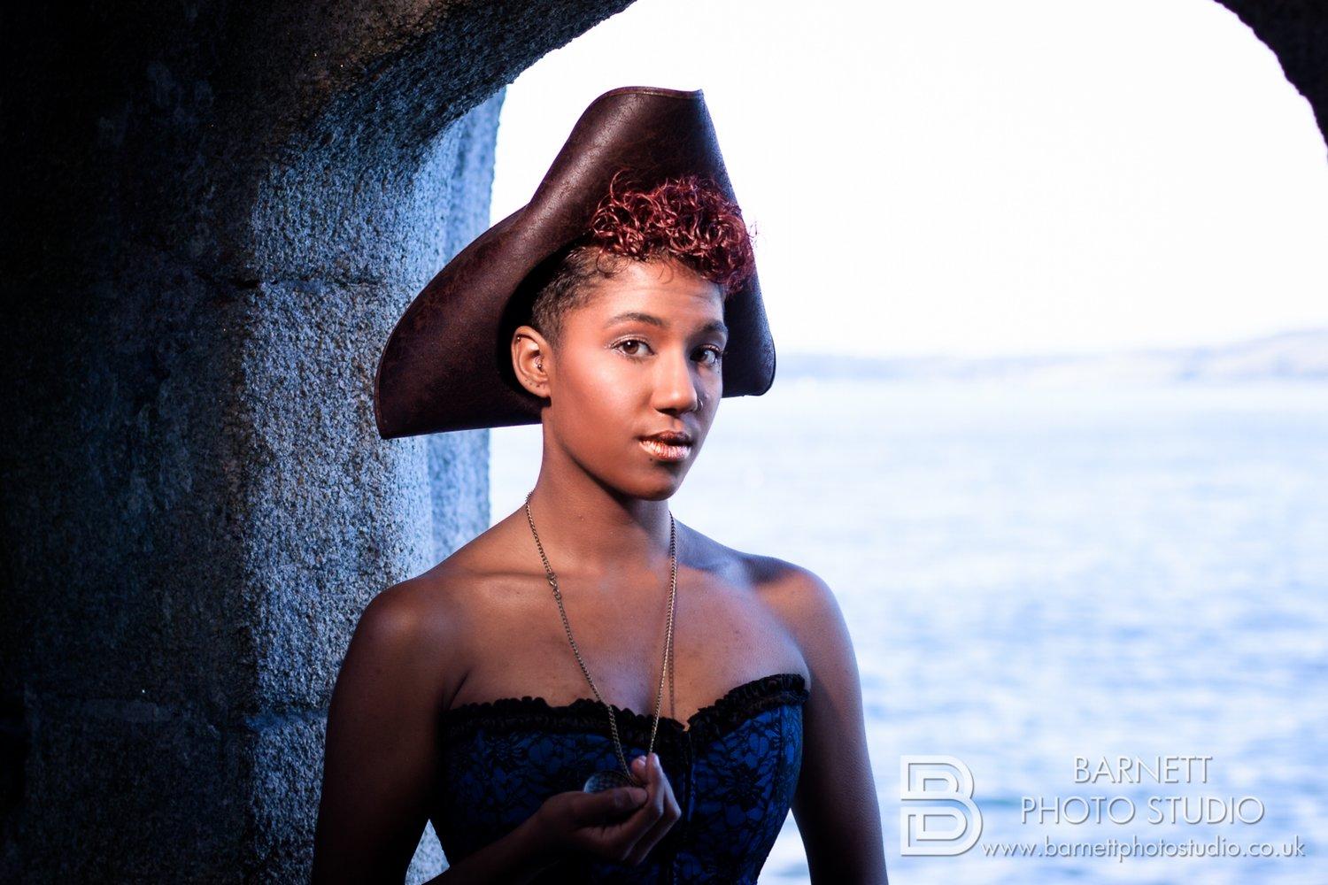 Rhyana standing in a stone archyway in a blue corset and pirate's tricorn hat, against an ocean backdrop.