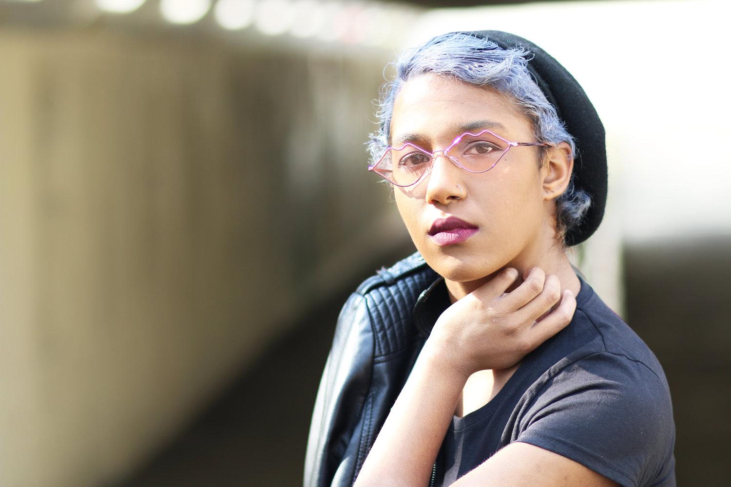 Rhyana looking towards camera with one hand on their neck, black leather jacket on their right shoulder.