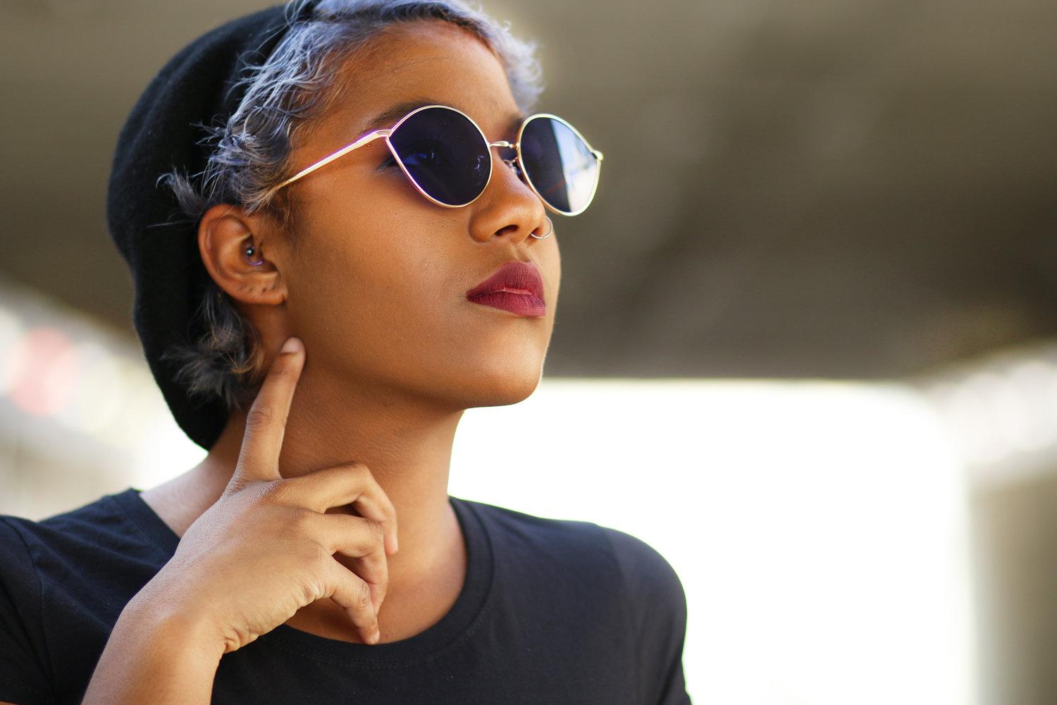 Rhyana looking towards the right with one finger on their neck, wearing sunglasses and a black beanie.