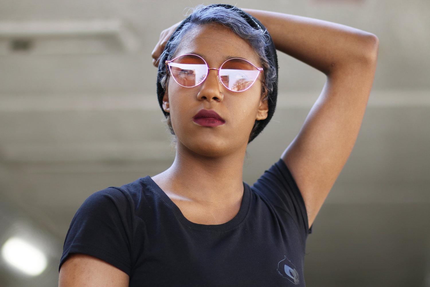 Rhyana in rose sunglasses and dark blue t-shirt, with one arm behind their head, looking towards the camera.