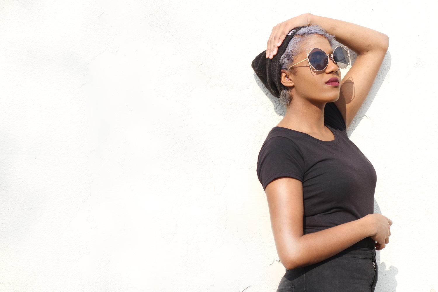 Rhyana against a bright white background, wearing a dark blue t-shirt, sunglasses and a black beanie.