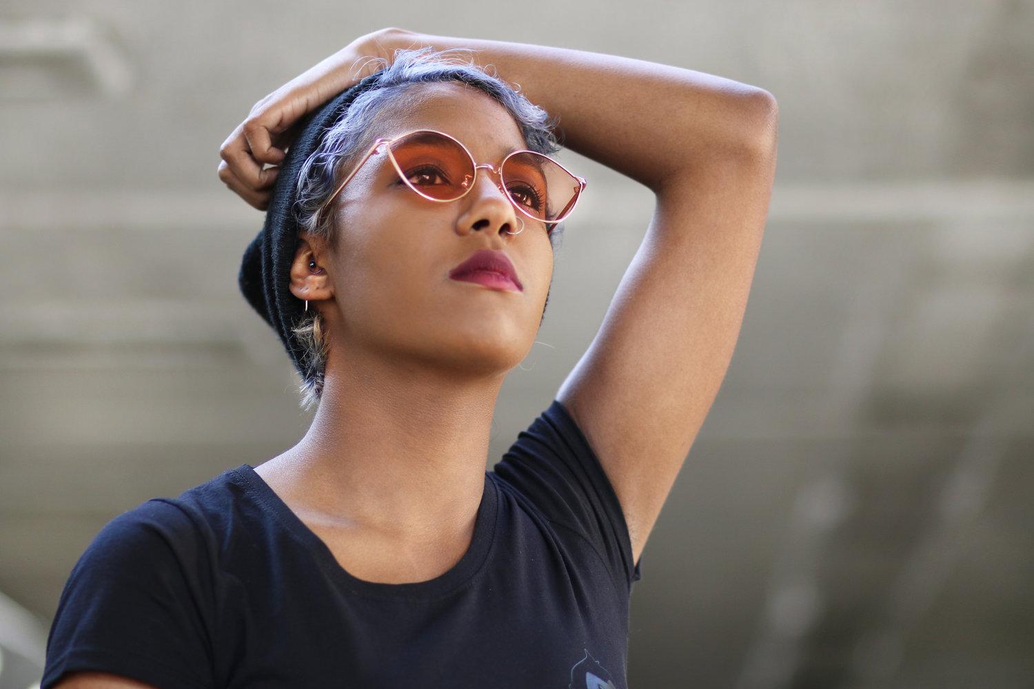 Rhyana with one hand behind their head, looking upwards off camera through rose sunglasses.