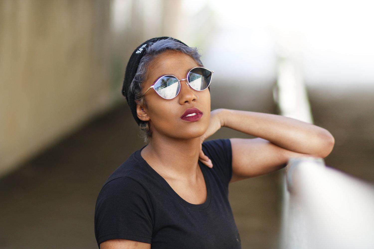 Rhyana leaning one arm against a railing, looking through mirrored sunglasses in a black t-shirt.