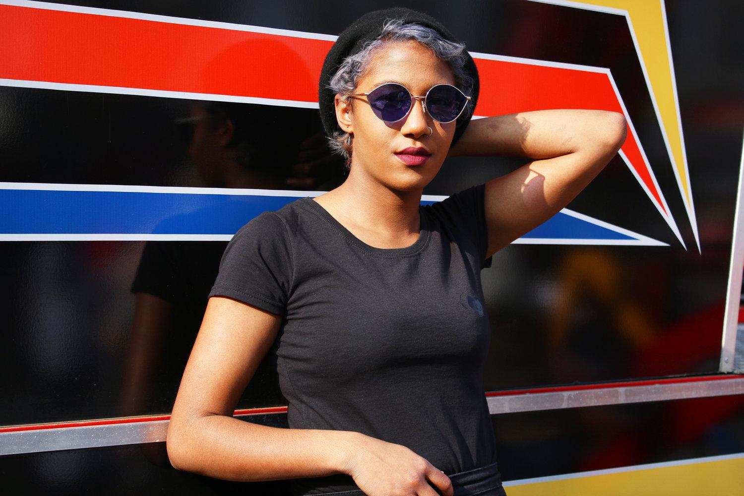 Rhyana leaning against a black, red, blue and yellow background, their hand behind their head.