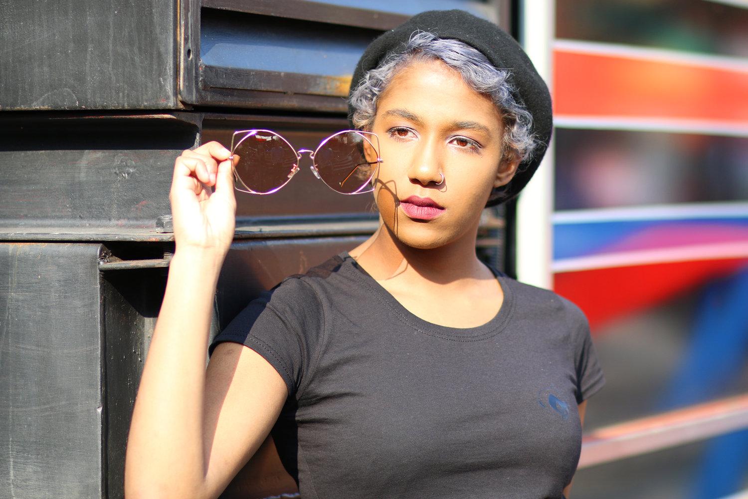 Rhyana looking at a pair of rose sunglasses, dressed in dark clothing.