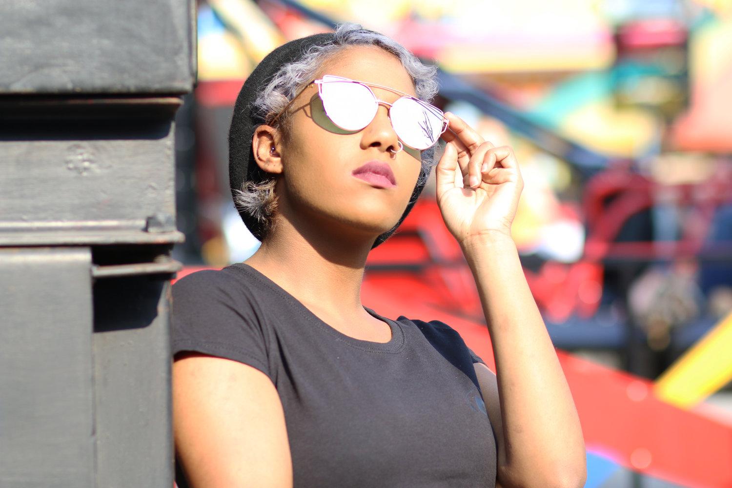 Rhyana leaning against a wall, looking upwards through reflective sunglasses.