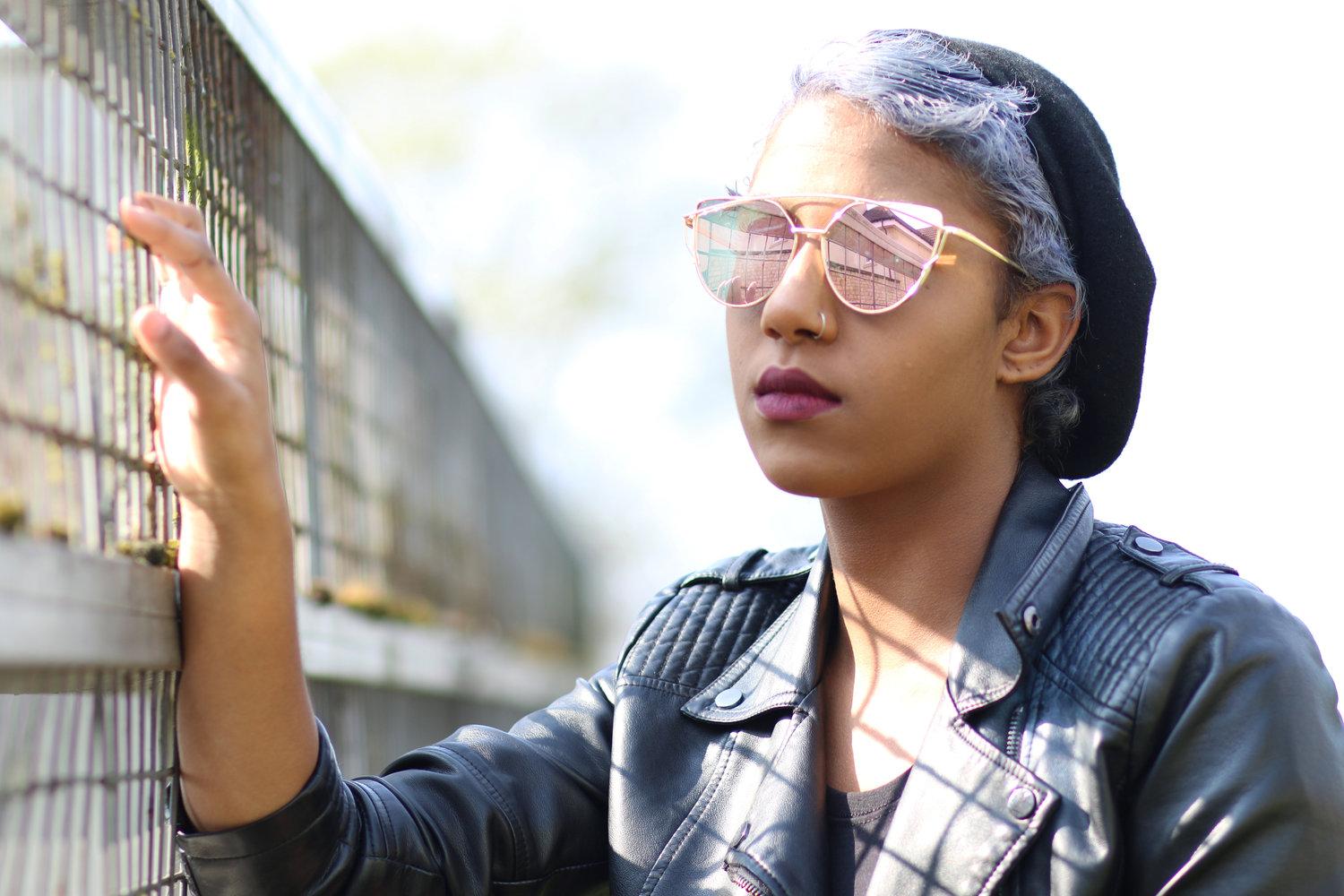 Rhyana with one hand on a rectangular fence in a black leather jacket and rose sunglasses.