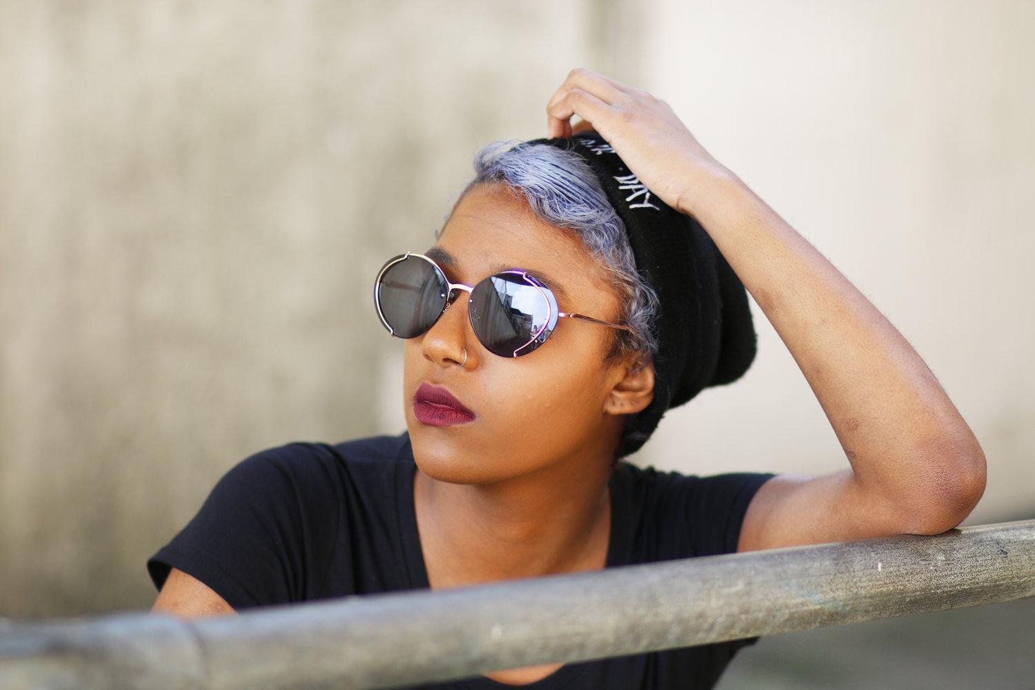 Rhyana leaning against a railing with one hand on their head, looking towards the right.