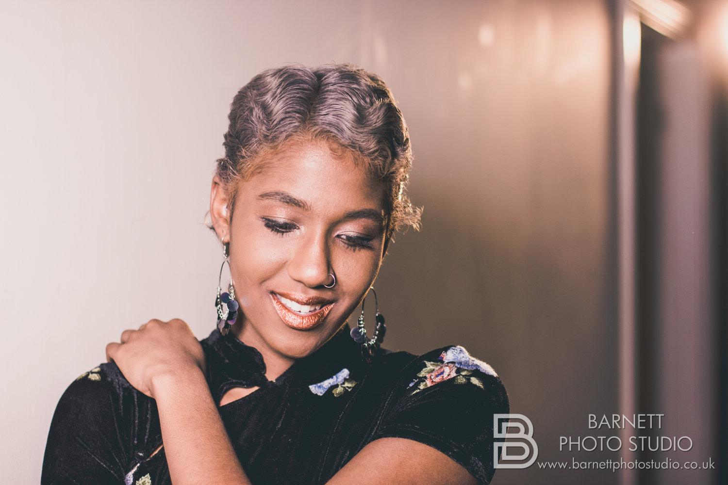 Rhyana smiling towards the ground in a black floral dress, one arm crossed over their other shoulder.