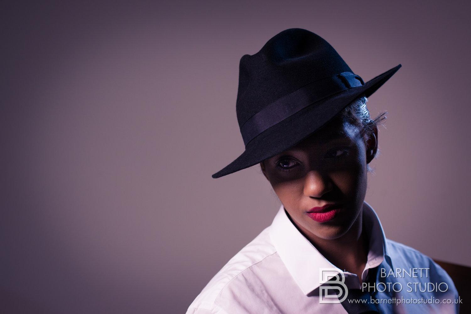 Rhyana in a wide-brimmed black hat and white shirt, looking off camera against a purple background.