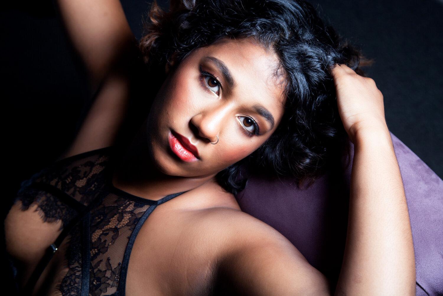 Rhyana in black lace lingerie leaning against a purple sofa, looking towards the camera.