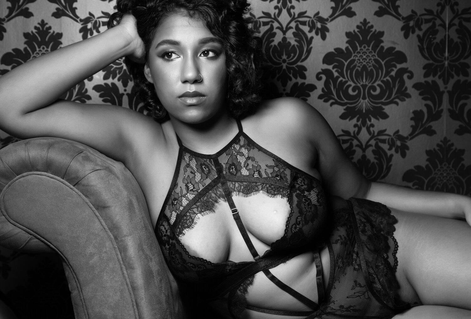 Rhyan in black and white, leaning against the arm of a sofa in lace lingerie, and gazing towards the right.