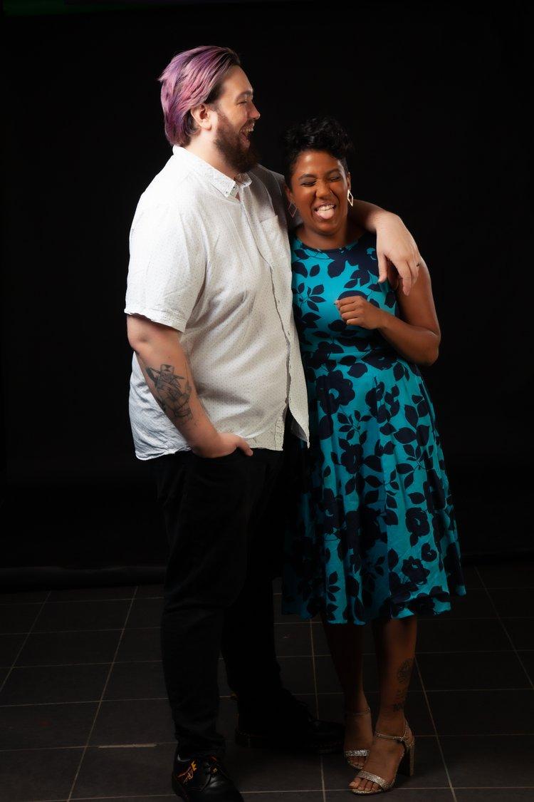Rhyana in a blue and black floral dress, laughing with partner Matt, who wears a white shirt and black trousers.