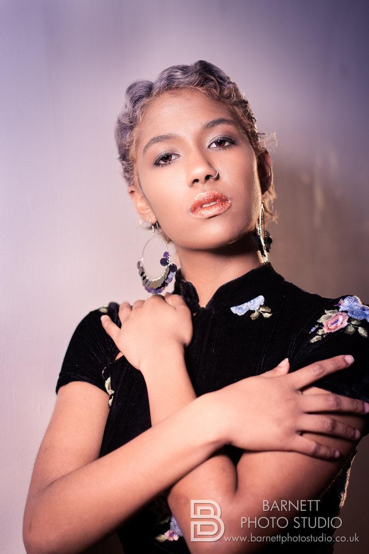 Rhyana wearing a black, purple and blue floral dress with arms crossed, looking towards camera against a purple background.
