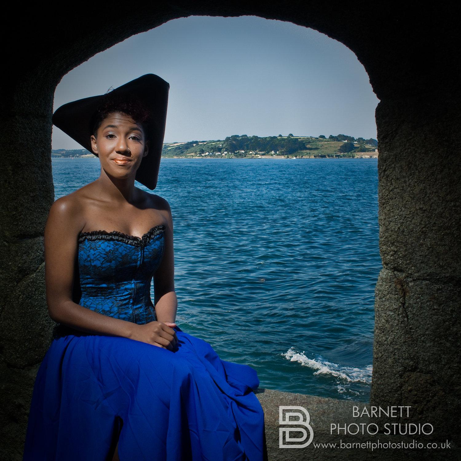 Rhyana sat in a stone archway in a blue and black corset with tricorn hat.
