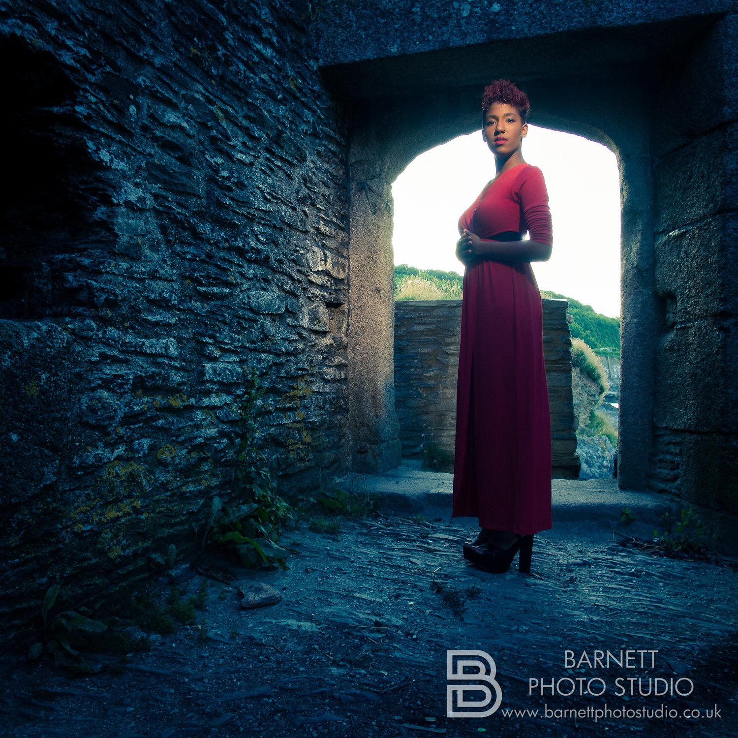 Rhyana in a red dress standing with hands on her waist in a stone corridor, ending in an archway.