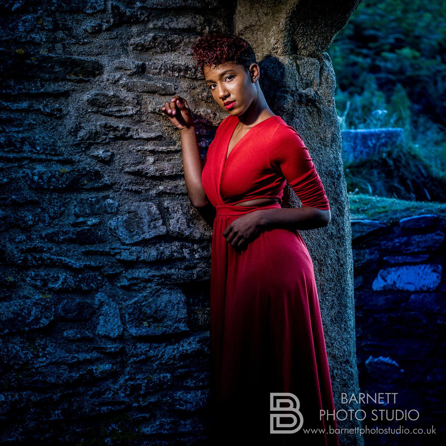 Rhyana leant against a cobbled stone wall in a red dress.