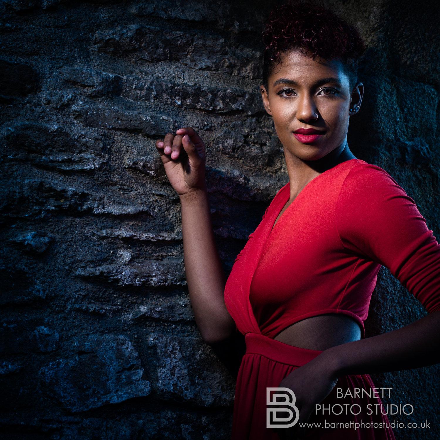Rhyana smiling with one arm against a cobbled stone wall, dressed in a red dress.