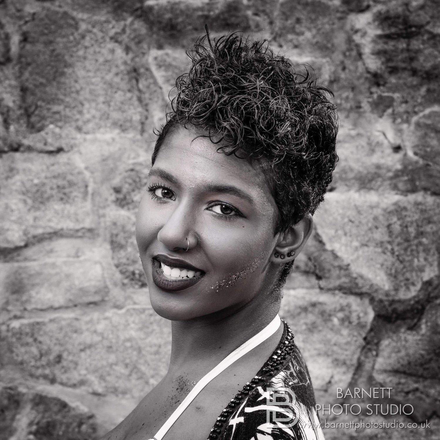 Rhyana in black and white, smiling towards camera in front of a cobblestone wall.