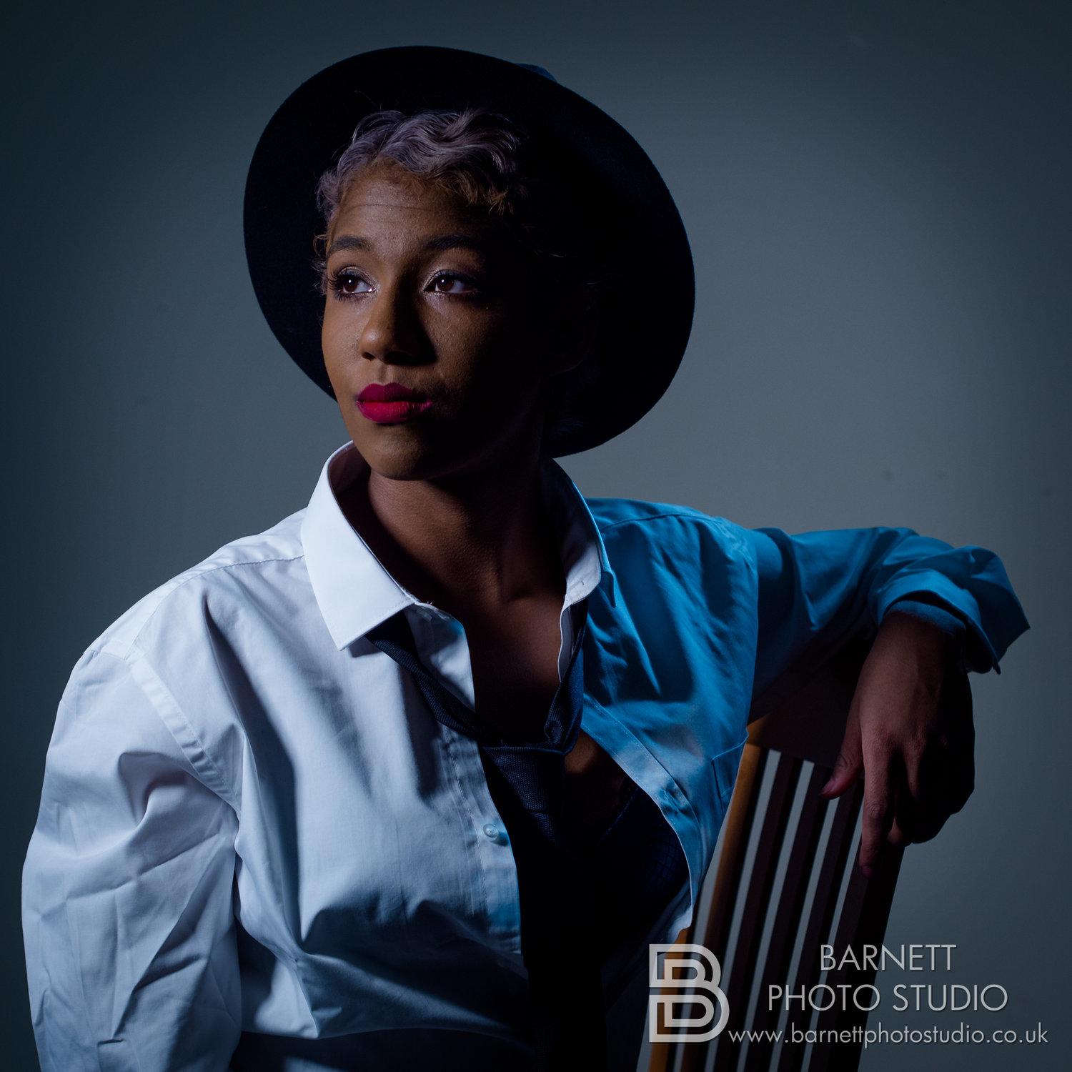 Rhyana in a wide-brimmed black hat and open white dress shirt, sat leaning against the back of a chair.