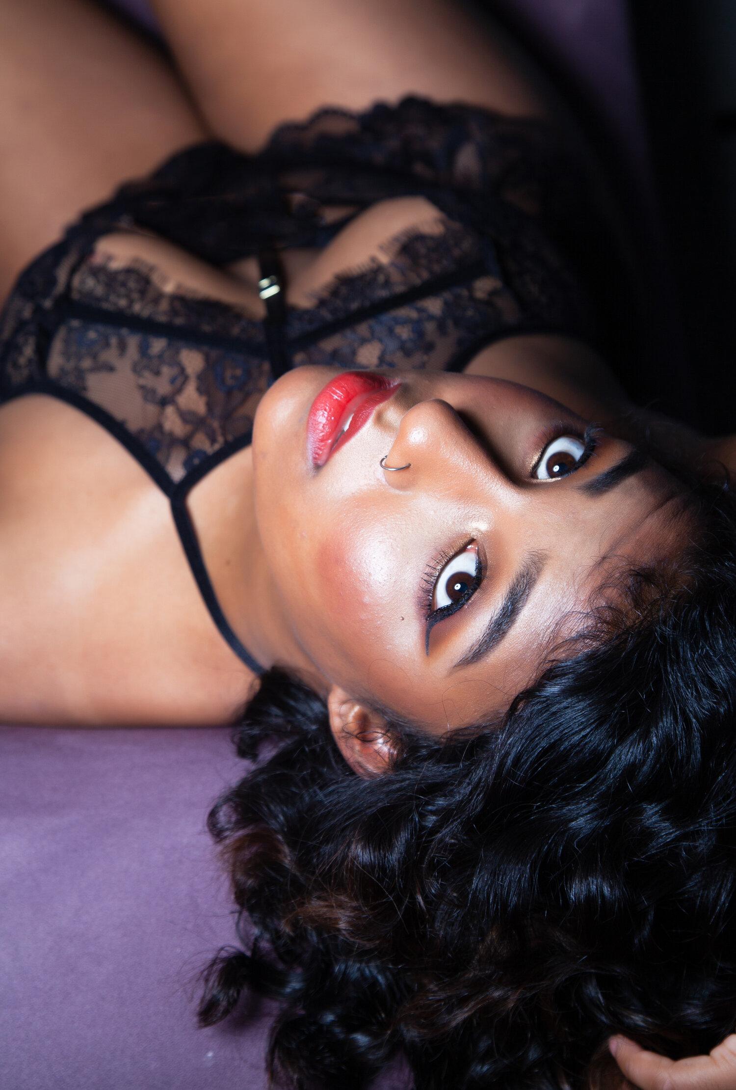 Rhyana in black lace lingerie, laying on a purple sofa and looking upside-down towards the camera.