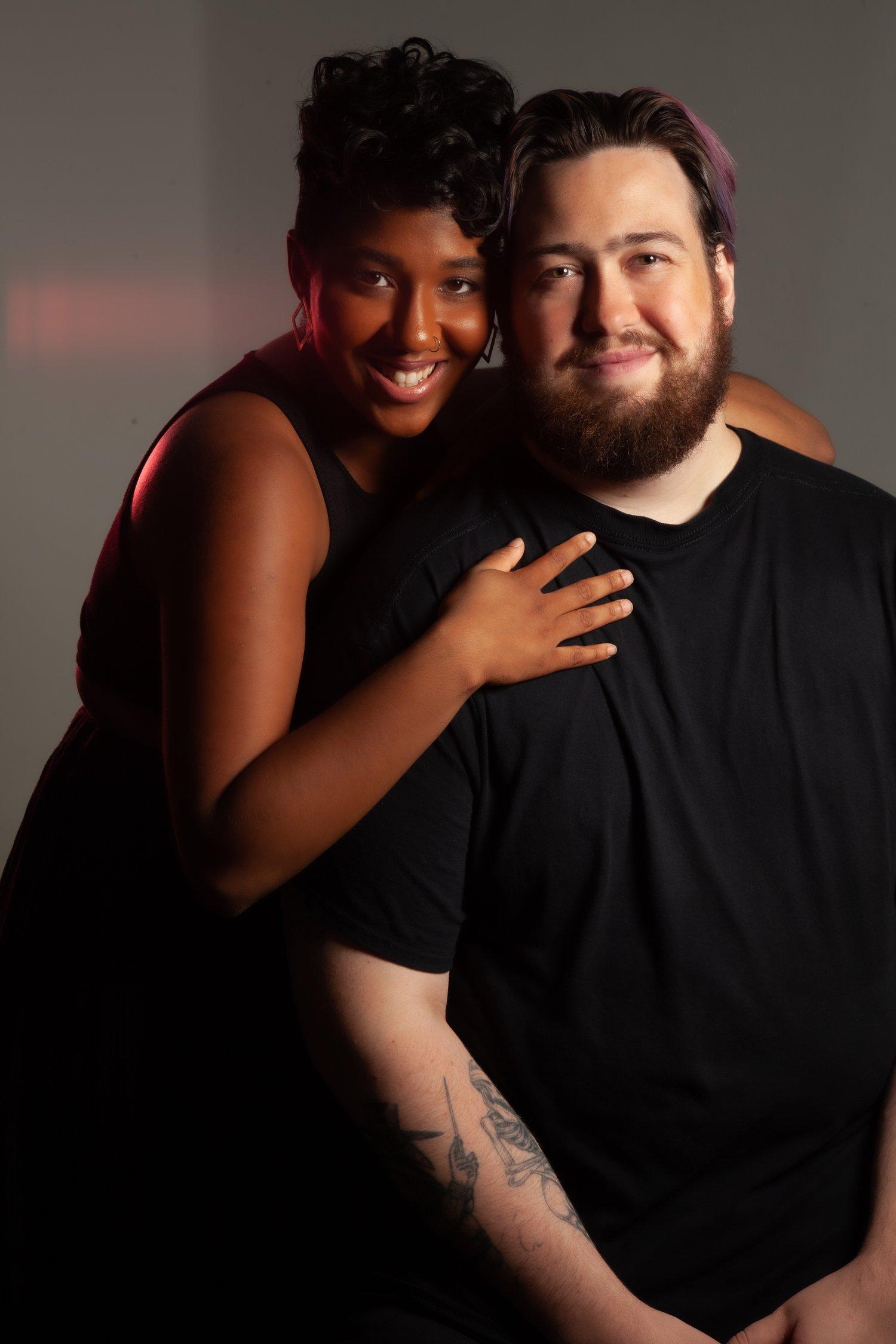 Matt sat in a black t-shirt, being embraced by Rhyana, who wears a black sleeveless dress.