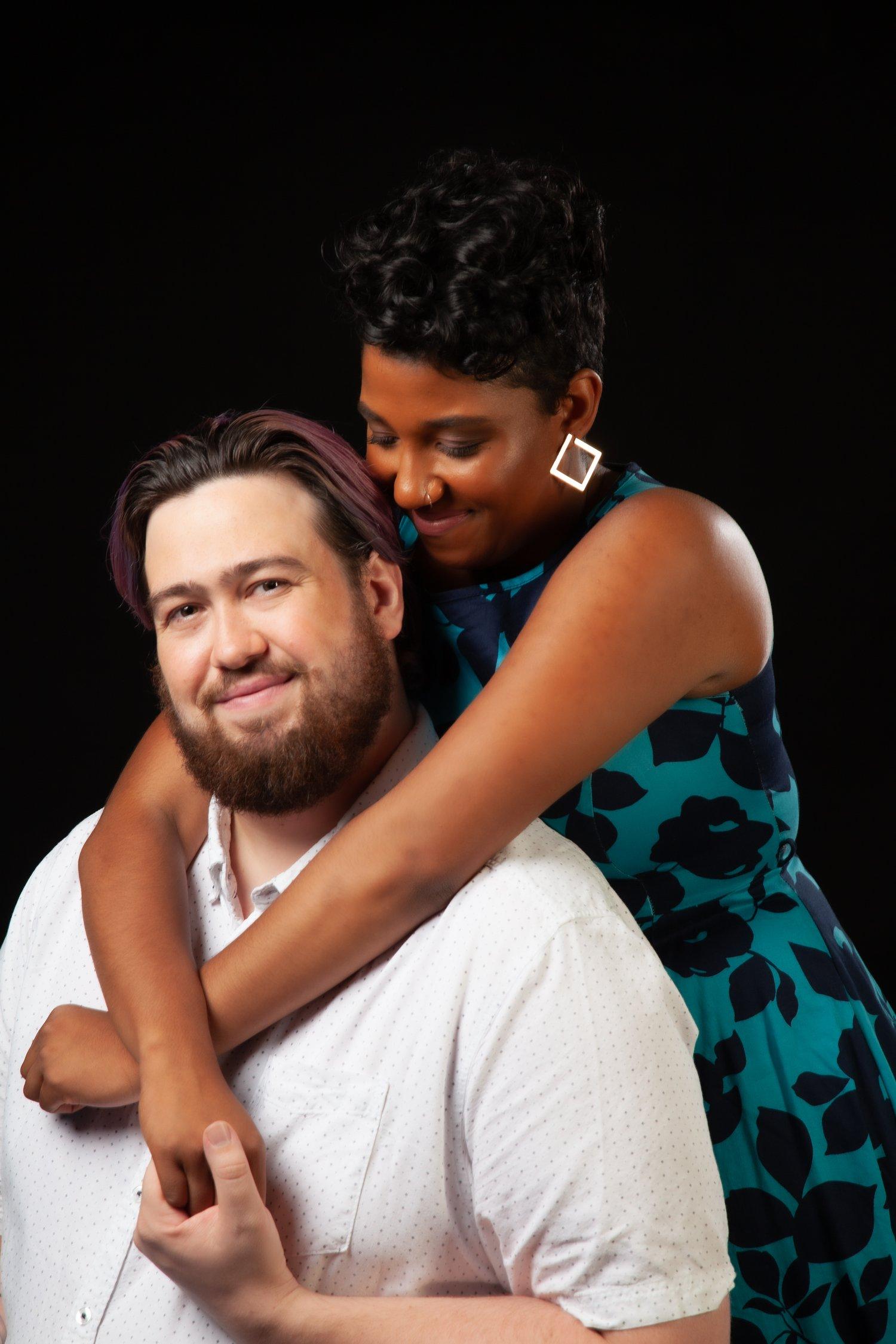 Rhyana embraces Matt from behind, who is sat in a white shirt and smiling towards the camera.