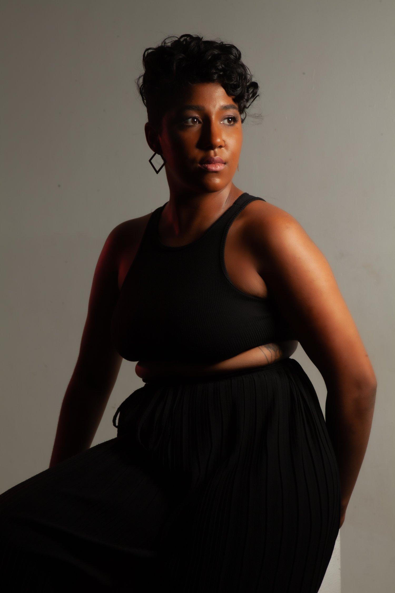 Rhyana looking off-camera, sat in a sleeveless black top and matching skirt.