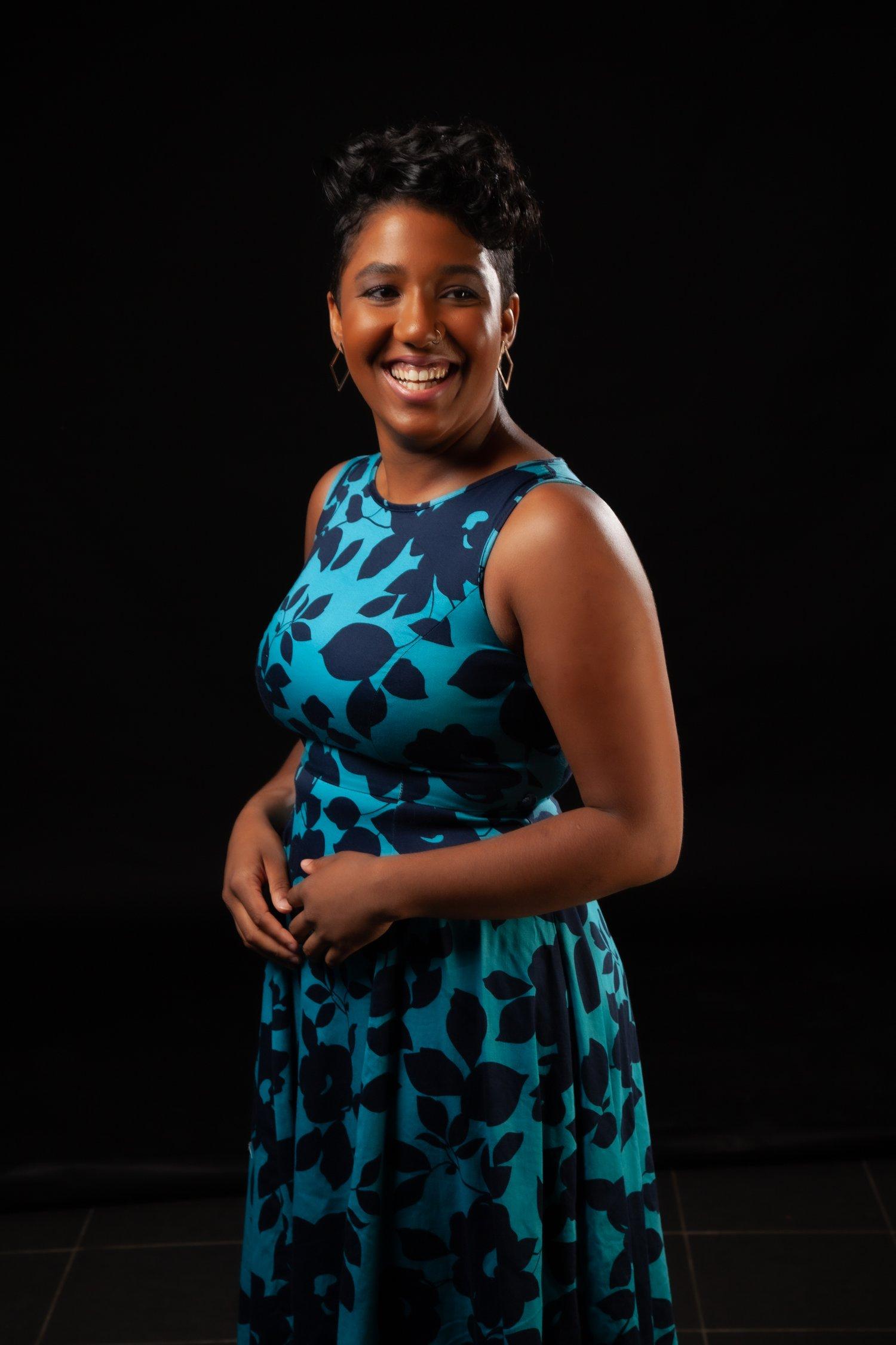 Rhyana in a blue and black floral dress stood smiling against a black background.