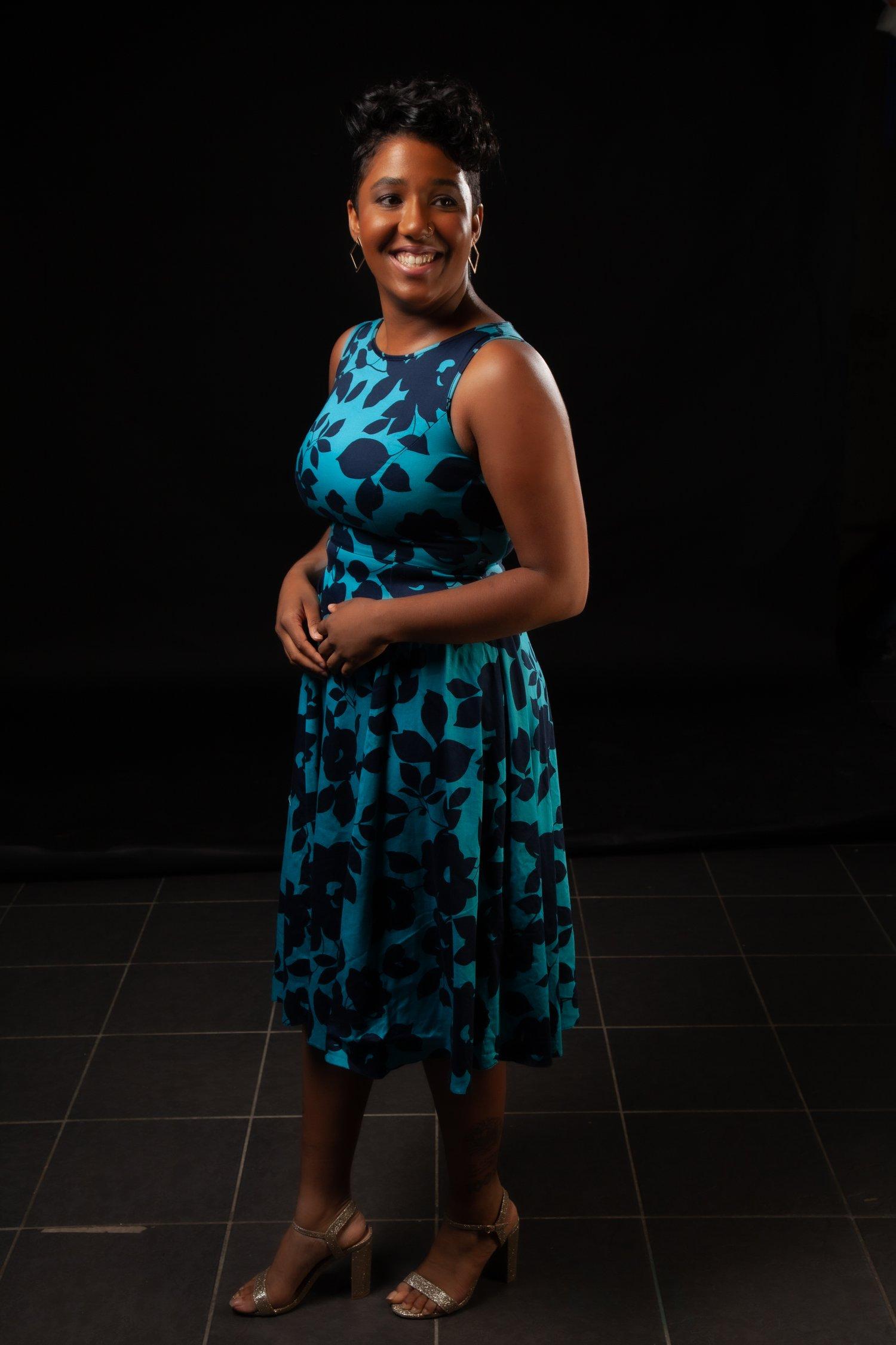 Rhyana in a blue and black dress and brown heels, stood smiling against a black tiled background.