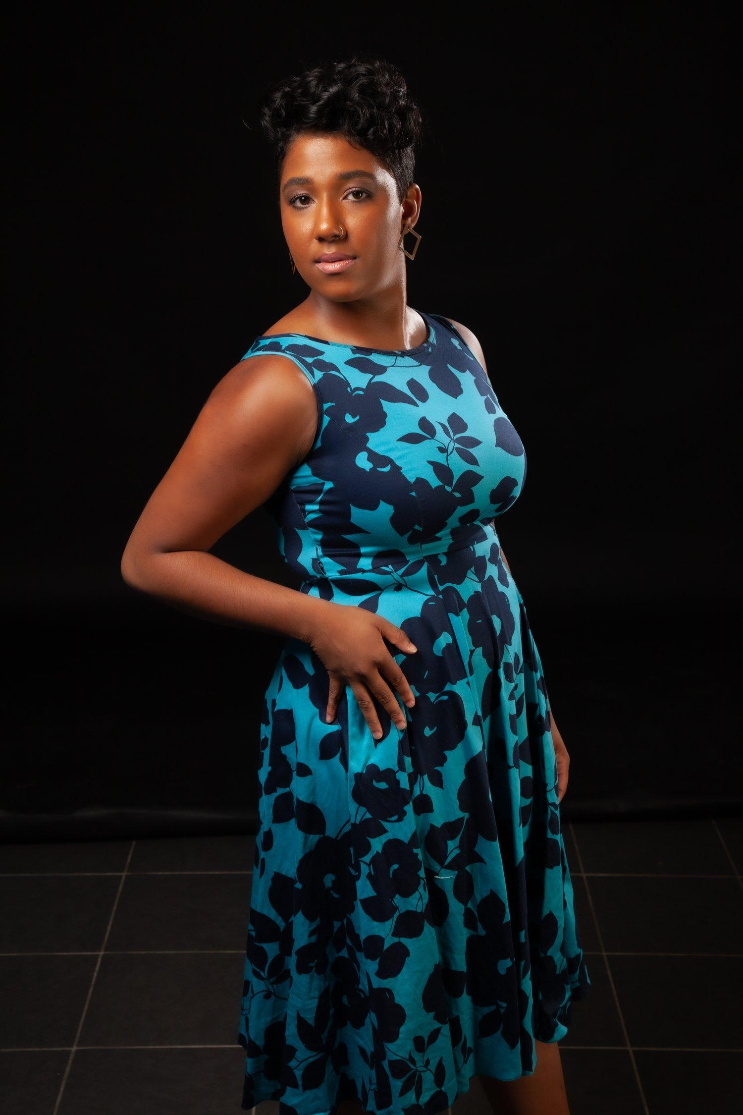 Rhyana with one hand on their hip, stood against a black background in a blue and black floral dress.