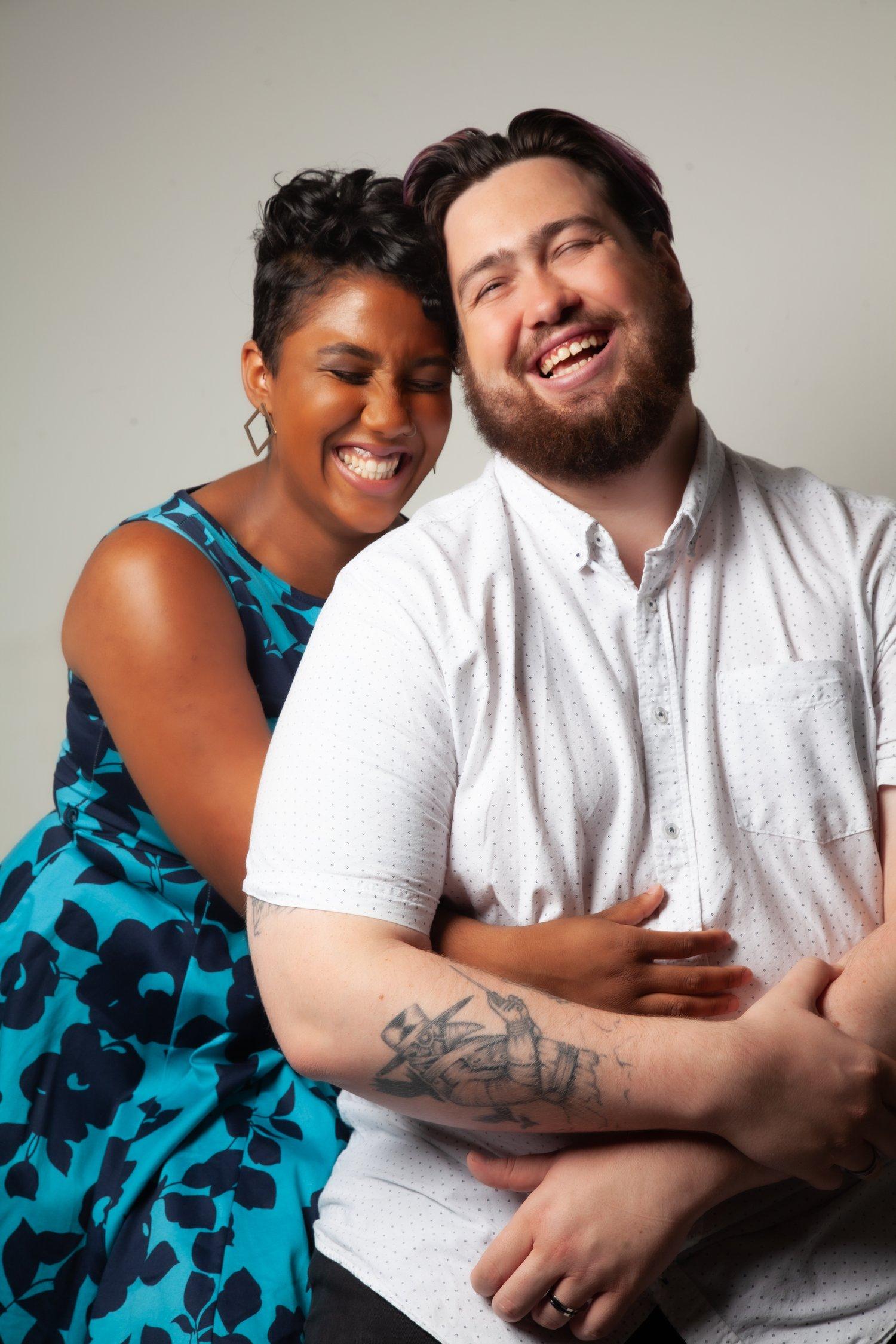 Matt sat in a white shirt while Rhyana embraces him, both mid laughter.