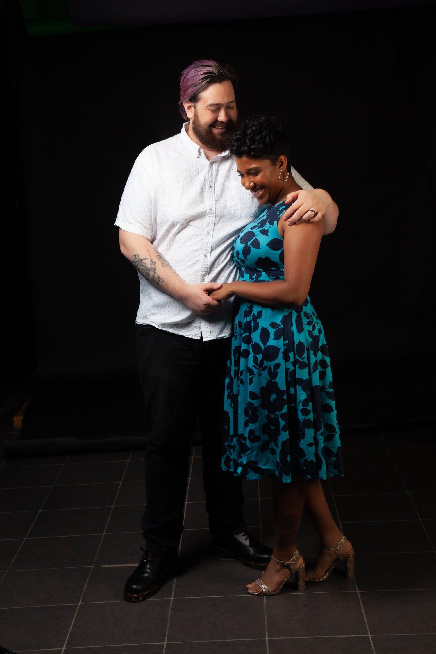 Rhyana and Matt embrace and smile together against a black background.