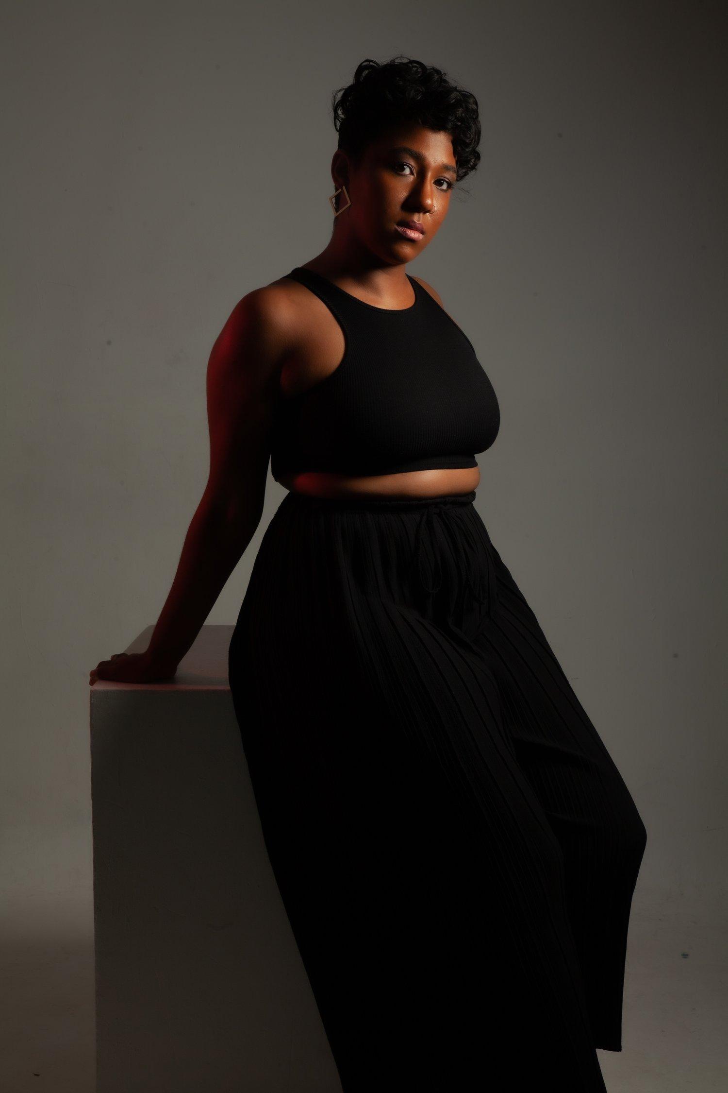 Rhyana leaning back in a black sleeveless top and matching skirt against a white background.