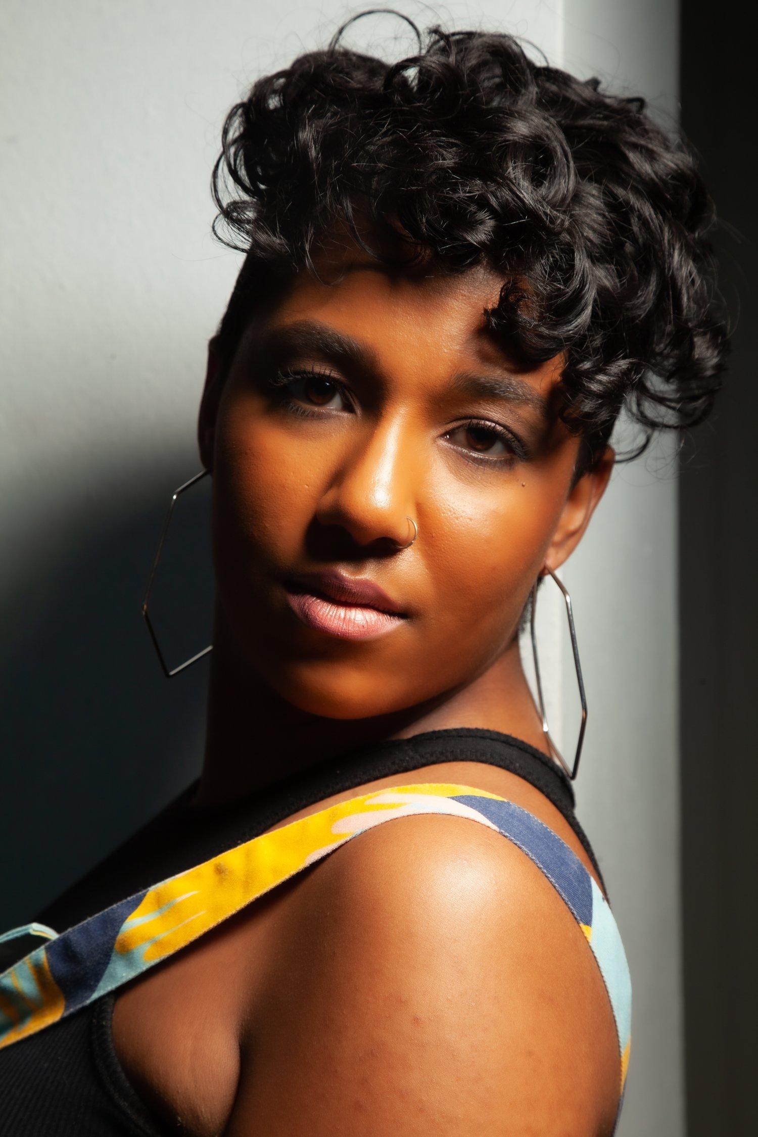 Close up of Rhyana looking towards the camera, wearing a yellow, white and blue dress and hexagonal earrings.