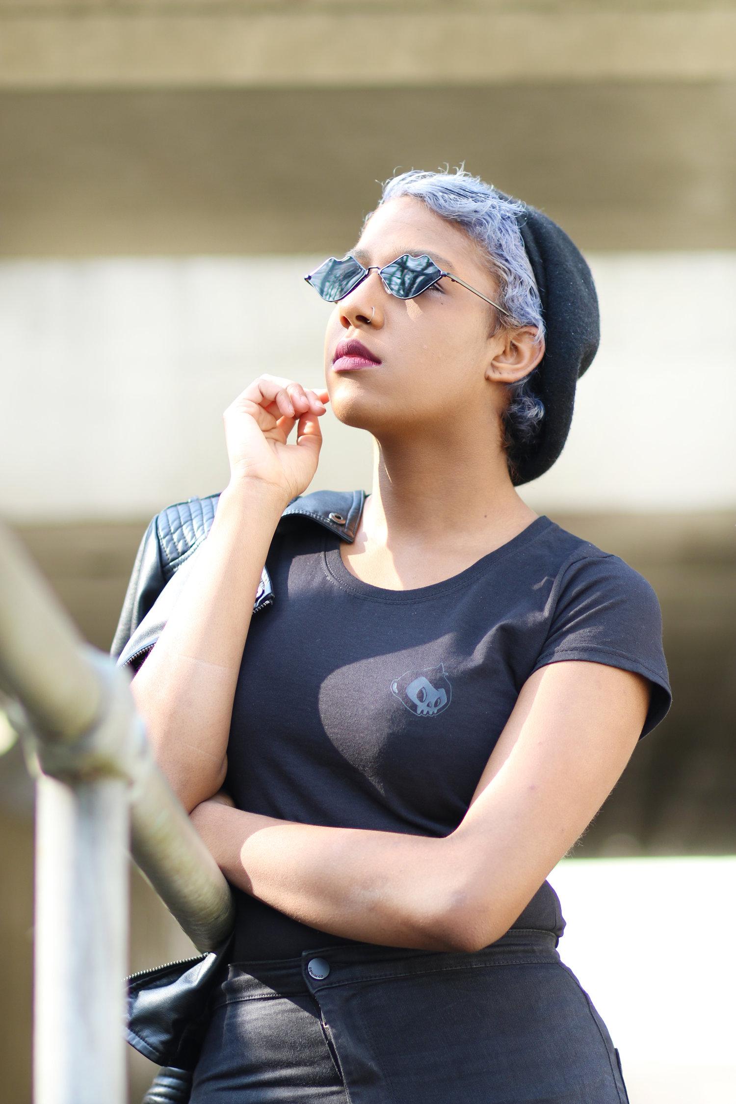 Rhyana in a dark blue t-shirt and dark jeans, looking through rose sunglasses towards the sky with their arms folded.
