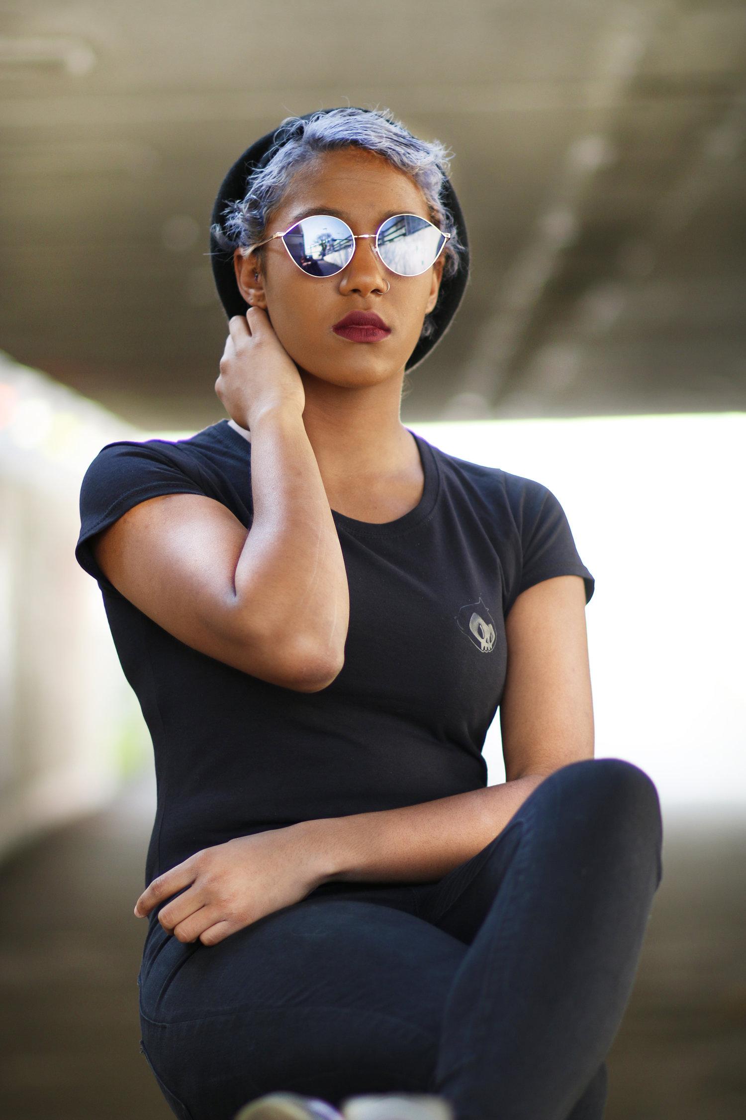 Rhyana sat in dark clothing and reflective sunglasses, with one hand on their neck and legs crossed.