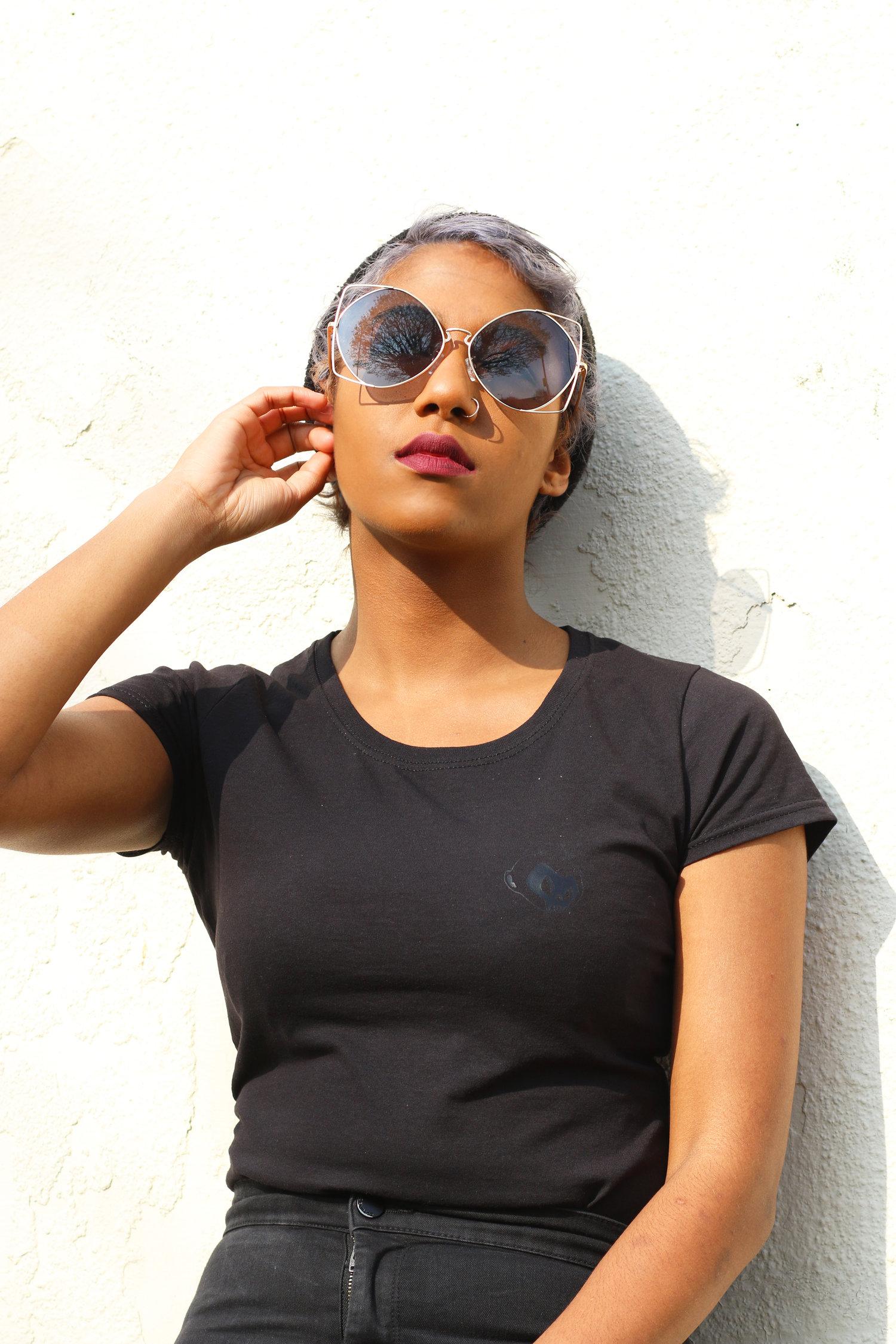 Rhyana leaning against a white wall in dark clothing, with one hand on their sunglass frames.