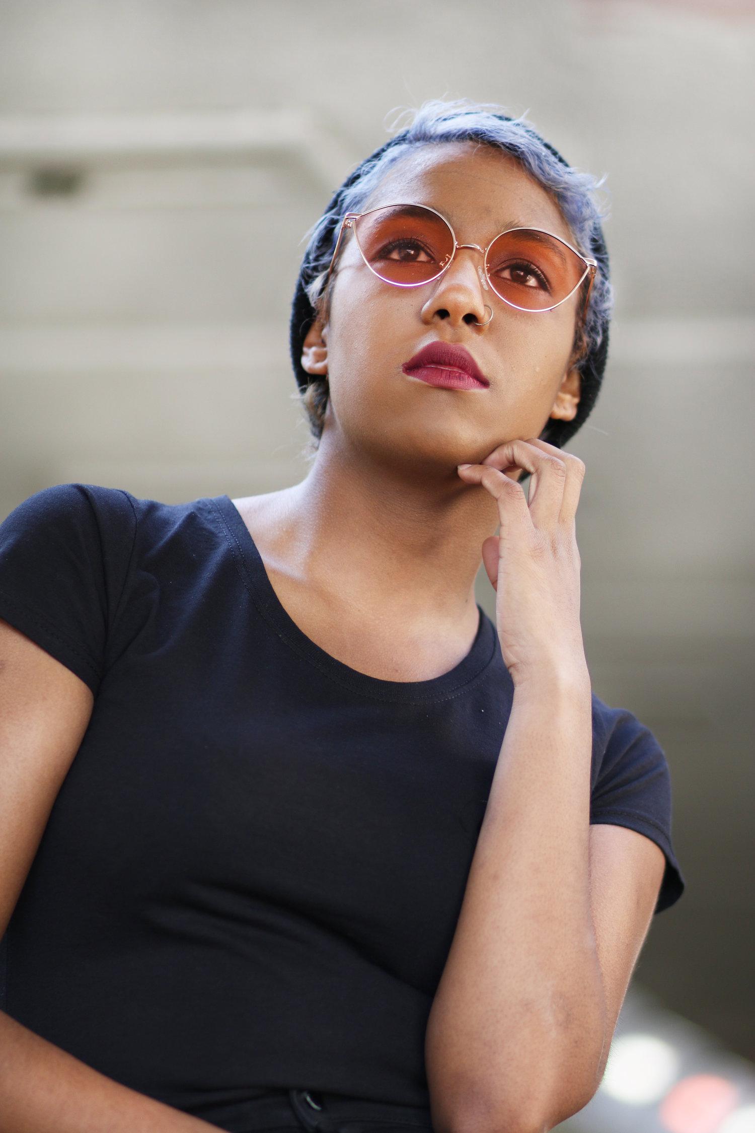 Rhyana leaning on one arm looking upwards through rose sunglasses.