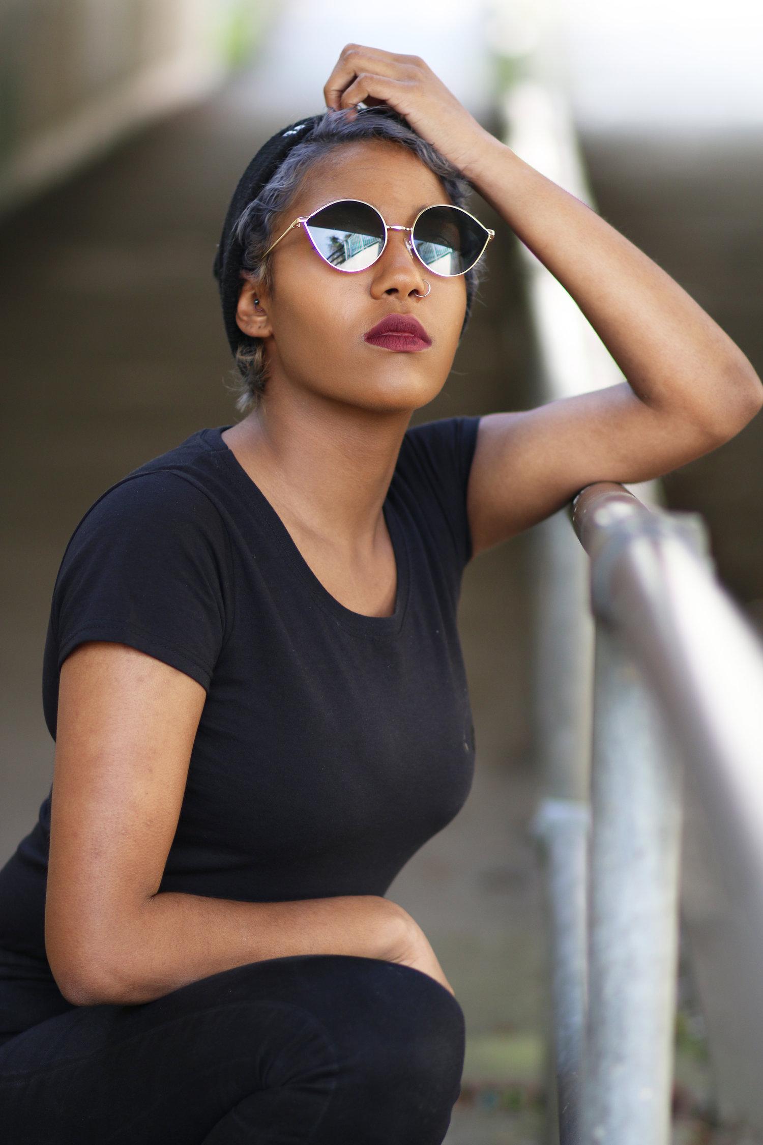 Rhyana kneeling with one arm leaning on a railing, hand on their head and the other on their knee.