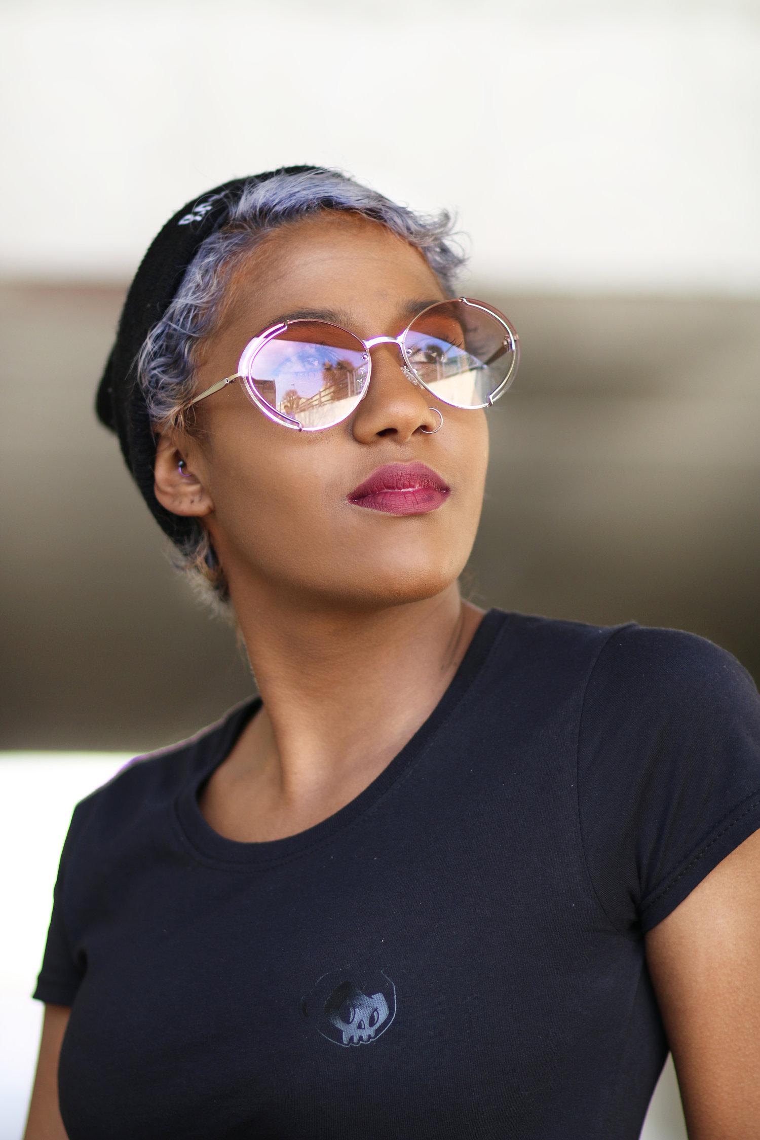 Rhyana in a dark blue t-shirt, looking off-camera through rose sunglasses.