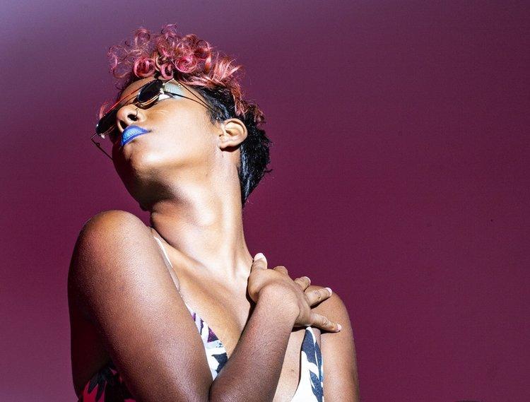 Rhyana in a white floral dress with pink hair and dark glasses, one hand against their chest, stood against a dark purple background.