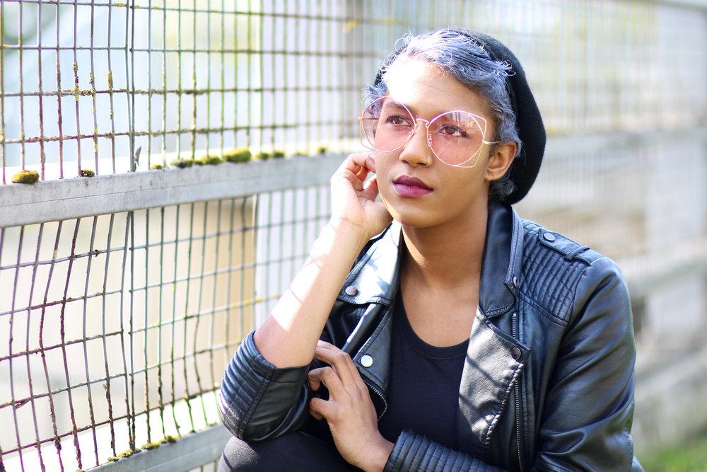 Rhyana in a black leather jacket and rose sunglasses, leaning against a fence on one elbow.